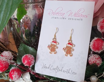 May include: Gold-tone gingerbread man earrings with red and white accents. The earrings are displayed on a white card with the text "Rebecca Williams JEWELLERY DESIGNER" and "Hand Crafted with Love". Festive holiday jewelry.