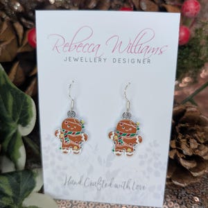 May include: A pair of gingerbread man earrings on a white card with the text "Rebecca Williams JEWELLERY DESIGNER" and "Hand Crafted with Love". The earrings are brown with white icing details, a green scarf, and silver hooks.