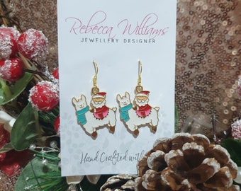 May include: Gold-tone earrings featuring white llamas wearing Santa hats and blue scarves. The earrings are on a white card with the text "Rebecca Williams JEWELLERY DESIGNER" and "Hand Crafted with". The background is a sparkly gold surface.