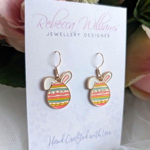 Puede incluir: Un par de pendientes dorados con forma de huevo de Pascua y orejas de conejo. Los huevos están decorados con rayas horizontales de colores. Los pendientes se exhiben en una tarjeta blanca con el texto "Rebecca Williams Jewellery Designer" y "Hand Crafted with Love."
