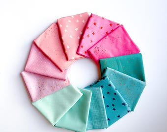 May include: A circular arrangement of twelve folded fabric squares in a gradient of colors ranging from light pink and coral to hot pink, teal, and bright blue. Each piece of fabric features distinct patterns including small stars and dots.