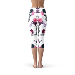May include: White capri leggings with a repeating pattern of black skulls and pink and red roses. The leggings have a floral and gothic design, perfect for a unique and stylish look. The capri length ends just below the knees.