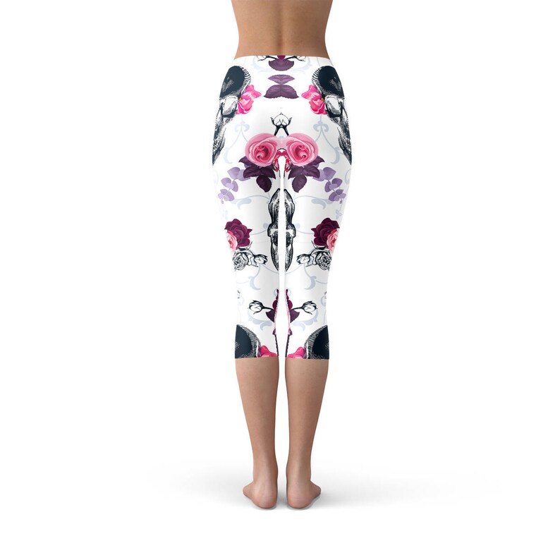 May include: White capri leggings with a repeating pattern of black skulls and pink and red roses. The leggings have a floral and gothic design, perfect for a unique and stylish look. The capri length ends just below the knees.