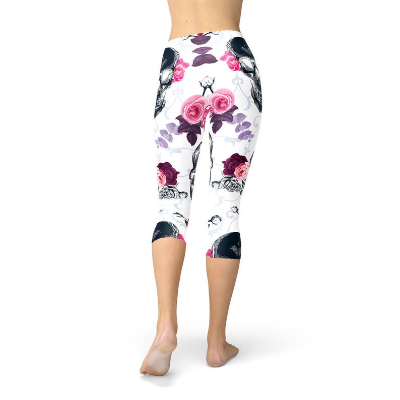 May include: White capri leggings with a repeating pattern of black skulls and pink and burgundy roses. The leggings have a floral design with purple accents. The model is wearing the leggings.