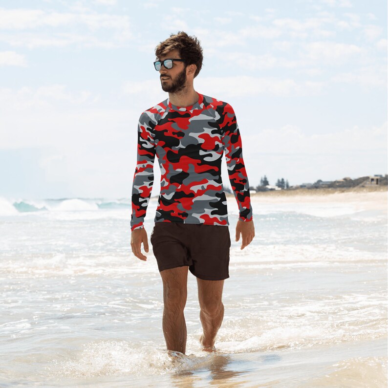 Pu&ograve; includere: Un uomo indossa una camicia a maniche lunghe mimetica rosso, nero e grigio e pantaloncini marroni scuri e cammina su una spiaggia.