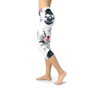May include: White capri leggings with a repeating pattern of black skulls, pink roses, and purple butterflies. The leggings have a high waist and a cropped length, suitable for various activities.