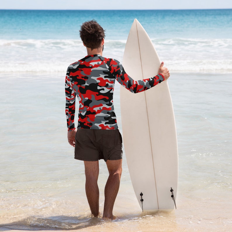 Pu&ograve; includere: Una persona indossa una maglietta a maniche lunghe mimetica rosso, nero e grigio e pantaloncini neri e si trova su una spiaggia tenendo una tavola da surf bianca.