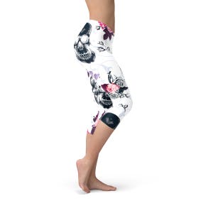 May include: White capri leggings with a black skull and floral design. The leggings feature a repeating pattern of skulls, roses, and other flowers in shades of pink, purple, and black. The leggings are made of a stretchy material.