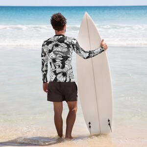 May include: A person wearing a black and white long-sleeved shirt with a floral and fish design, holding a white surfboard on a beach.