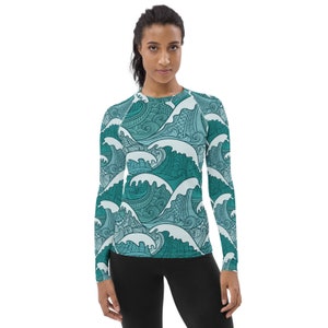 May include: A long-sleeved rash guard shirt featuring a teal and white wave pattern. The shirt has a crew neck and is made of a stretchy material. The wave design is detailed with swirling lines and patterns. The model is wearing black leggings.
