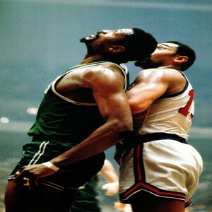 May include: A vintage photograph captures two basketball players in a moment of intense competition. One player wears a green and white uniform, while the other is in a white and red uniform. The players are embracing, their faces close together.