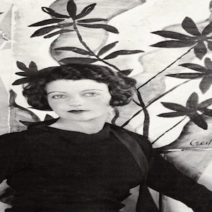 May include: A black and white portrait of a woman with dark hair and dark lipstick. She is wearing a dark top. Behind her is a painting of large, stylized black flowers and foliage. The artist's signature, "Cecil," is visible.