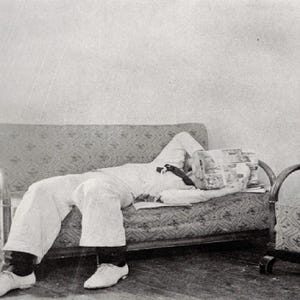 May include: A vintage black and white photograph depicts a person reclining on a patterned sofa, with a newspaper covering their face. The person is wearing white clothing and white shoes. An armchair is to the right.