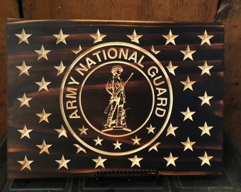 May include: A wooden American flag with the Army National Guard seal in the center. The seal is a circle with the words "Army National Guard" around the outside and a soldier with a rifle in the center. The flag is dark brown with light brown stars.