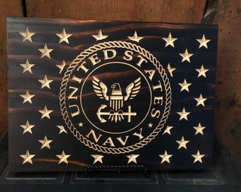 May include: A wooden plaque with a dark brown background and a gold-colored United States Navy seal. The seal features an eagle with wings spread, an anchor, and a cross. The seal is surrounded by a rope design and the words "United States Navy". The plaque is decorated with 48 gold-colored stars.