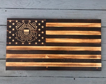 May include: A wooden American flag with a distressed finish. The flag features the United States Coast Guard emblem in the upper left corner. The emblem is a gold color and features a shield with the words "United States Coast Guard 1790" around it.