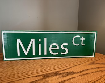 May include: A green street sign with white lettering that reads "Miles Ct". The sign has a white border and is made of wood. The sign is displayed on a wooden surface.