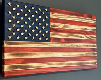 May include: Wooden American flag with a blue canton featuring 50 wooden stars. The flag's stripes alternate between red and natural wood tones. The flag is mounted on a wall, showcasing a rustic, patriotic design.