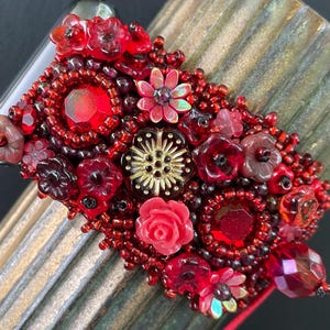 以下が含まれることがあります： 赤いビーズと金のビーズでできた、花柄のブレスレット。赤いビーズ、赤い花、金のアクセントでできています。