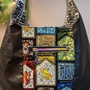 May include: Black leather tote bag with a colorful fabric lining featuring stained glass style Hogwarts house crests. The fabric lining includes the crests for Gryffindor, Hufflepuff, Slytherin, and Ravenclaw. The bag has a zipper closure and a personalized name tag that reads "Mioni".