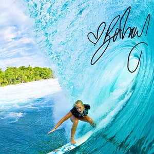 May include: A woman surfs a large wave in a tropical location. The wave is a bright turquoise blue and the water is a deep blue. The woman is wearing a black wetsuit and a red bikini bottom. She is holding a surfboard with a blue and white design. The sky is blue with white clouds. The word "Bohdi" is written in black cursive on the wave.
