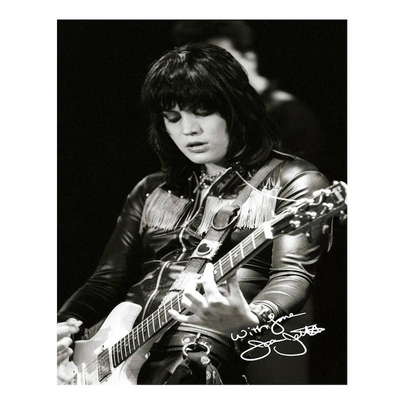 May include: Black and white photo of a woman playing a black electric guitar with a fringe detail on the jacket. The signature "With love Joan Jett" is written in white ink in the bottom right corner of the photo.