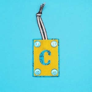 May include: A yellow felt tag with a blue letter C on it. The tag has four blue buttons around the edges and a black and white string attached to the top.