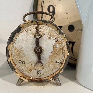 May include: A round, vintage-style metal timer with a distressed, cream-colored face featuring black numbers 5, 10, 15, and 20. The timer has a dark, rusted metal frame, a single central pointer, and two small metal feet at the base for support.