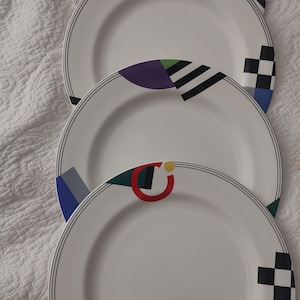May include: A set of four white ceramic dinner plates arranged in a vertical stack. Each plate features a geometric pattern along the rim, including black and white checkerboard squares, solid purple, blue, and green shapes, and a red circular arc.