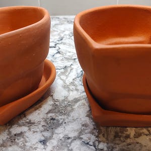 May include: Two terracotta plant pots with matching heart-shaped saucers. Each pot features a distinct heart-shaped rim and a matte, earthy orange finish. The pots are empty and designed with a wide opening, providing a simple structure.