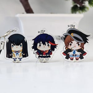 Op de afbeelding: Drie sleutelhangers met anime-personages. Elke sleutelhanger heeft een ander personage met uniek haar en outfits. De sleutelhangers zijn gemaakt van helder acryl en hebben zilverkleurige metalen sluitingen.