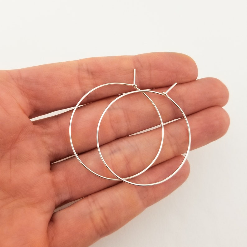 May include: A pair of silver hoop earrings. The hoops are thin and delicate, and they are shown on a hand.