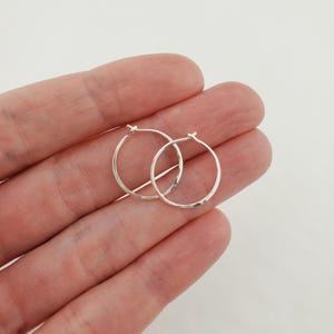 May include: Two small, silver hoop earrings. The earrings are round and have a hammered texture. The hoops are held in a hand against a white background. The earrings are a simple, minimalist design.