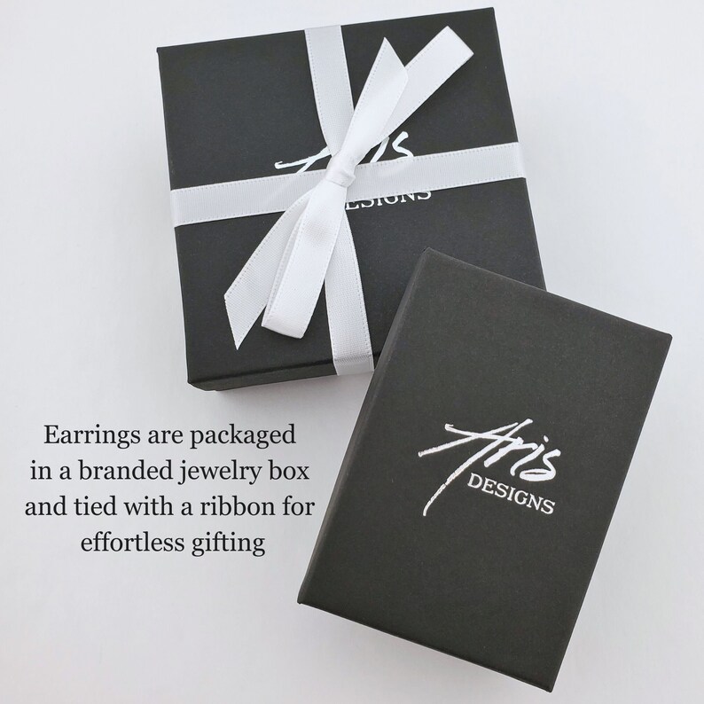 Earrings are packaged in a matte black jewelry box with Aris Designs logo stamped in metallic silver and tied with a white ribbon.