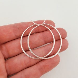 May include: A pair of silver hoop earrings with a hammered texture. The earrings are circular and have a simple, elegant design. The hoops are held in a hand against a white background.