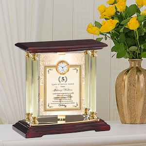 May include: A mahogany and gold-tone clock with a glass front displaying a certificate for 5 years of service award presented to Johnny Watkins. The certificate has a gold border and a gold seal with a laurel wreath. The clock is on a mahogany base with gold accents.