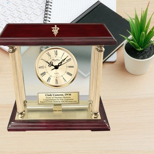 May include: A mahogany and gold desk clock with a glass front and a gold-toned face. The clock has a personalized plaque that reads "Cindy Cameron, DVM Doctor of Veterinary Medicine Congratulations On Your Graduation Engraved Desk Clocks by AllGames.com".