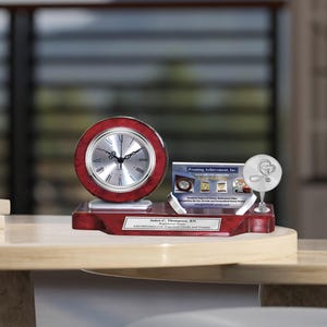 May include: A mahogany desk clock with a silver face and hands, a silver plaque with the text "Judy C. Thompson, RN Registered Nurse", and a silver plaque with the text "Framing Achievement, Inc." and a stethoscope and heart graphic.