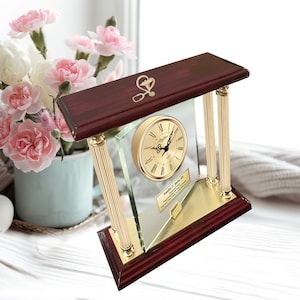 May include: A mahogany wood desk clock with gold accents and a glass face. The clock has a Roman numeral dial and a gold-toned stethoscope and heart symbol on the top.