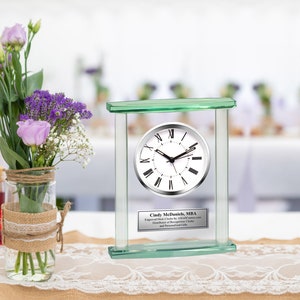 May include: A glass clock with a silver face and Roman numeral markings. The clock is framed in a clear glass rectangle with a green top and bottom. The clock is on a table with a lace tablecloth and two vases with purple flowers.