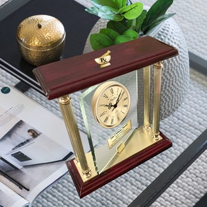 May include: A mahogany wood clock with gold accents and a glass face. The clock has a Roman numeral clock face and a gold plaque with the text "John S. Williams Pharmacy" and a mortar and pestle symbol.