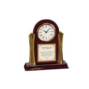 May include: A mahogany wood clock with gold accents and a white face. The clock has a plaque with the text "John Smith, MD Doctor of Medicine".