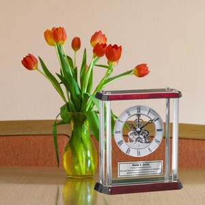 May include: A silver and glass clock with a skeletonized movement and a personalized plaque that reads "Wade E. Smith 5 Years of Dedicated Service We truly thank you for your hard work and dedication. AllGiftFrames.com Clocks and Frames". The clock is on a wooden table with a vase of orange tulips in the background.