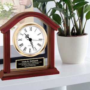 May include: A mahogany wood desk clock with a gold-toned face and Roman numeral hour markers. The clock is in a house-shaped frame with a clear glass front. A personalized plaque with the name "Cindy K. McDaniels" is attached to the base.