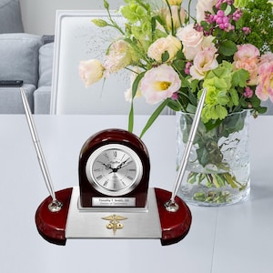 May include: A mahogany desk clock with a silver face and black hands. The clock is on a silver base with a gold medical symbol and the inscription "Timothy T. Smith, OD Doctor of Optometry". Two silver pens are in holders on either side of the clock.
