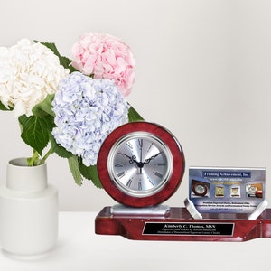 May include: A mahogany desk clock with a silver face and black hands. The clock is on a base with a silver plaque that reads "Kimberly C. Thomas, MSN Executive Director & Personalized Engraved Luxury Clocks Distributor of Personalized Engraved Luxury Clocks". A business card holder with a clear acrylic frame is to the right of the clock. The business card reads "Framing Achievement, Inc. www.framingachievement.com Premium Engraved Clocks, Retirement Gifts, Recognition Awards and Personalized Picture Frames". A bouquet of white, pink, and blue hydrangeas is in a white vase to the left of the clock.