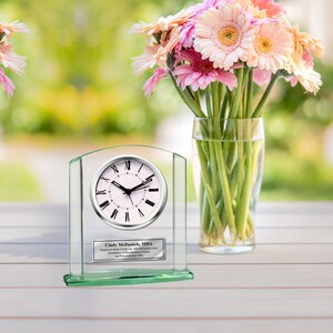 May include: A glass clock with a silver face and black Roman numeral hour markers. The clock is on a green glass base with an engraved plaque that reads "Cindy McDaniels, MBA, Engraved Desk Clocks by AllGiftFrames.com, Distributor of Recognition Clocks and Personalized Gifts".