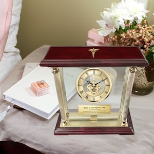 May include: A mahogany and gold desk clock with a glass front and a gold-toned Roman numeral clock face. The clock has a gold-toned medical symbol on the top and a personalized inscription on the bottom that reads "Kylie T. Thompson, Esq. Attorney at Law Congratulations on Your Graduation Engraved Desk Clocks by AllGiftFrames.com".