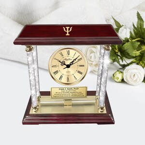May include: A gold desk clock with a mahogany base and crystal pillars. The clock face has Roman numerals and the words "The Allegiance" and "Quartz". A gold plaque with the text "Cindy J. Smith, PsyD Enjoy the gift of time We are so incredibly thankful for your 10 years of service at St. Thomas Hospital Engraved Desk Clocks by AlGiftFrames.com" is attached to the base.