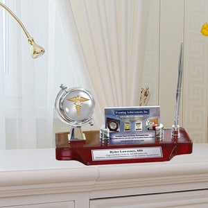 May include: A silver and gold desk set with a globe, pen holder, and a card that reads "Framing Achievement, Inc." The desk set is on a wooden base with the name "Ryder Lawrence, MD" engraved on it.
